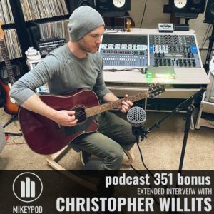 Christopher Willits is sitting in a recording studio with a guitar in his lap. He is wearing green pants, a green shirt, and a grey beanie.
