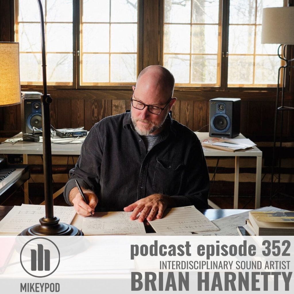 Brian Harnetty sits at a desk making writing with a pen on a stack of paper. There are other stacks of paper on the dest with handwritten music. Brian is a white male with a shaved head and a beard. He is wearing dark framed glasses and a long sleeved button down black shirt.