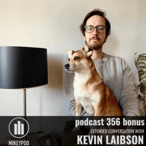 A man with glasses and facial hair holds a tan and white chihuahua against his chest, standing in front of a white wall with a black lamp and potted plant. MikeyPod logo and text overlay for podcast 356 bonus episode.