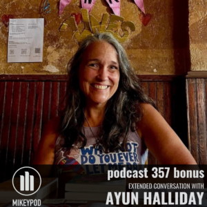 A smiling woman with long dark brown and gray hair sits in a dim, brown-toned bar or café, wearing a sleeveless graphic shirt. The wall behind her is textured with papers taped to it and wooden wainscotting below. The lower portion of the image contains overlaid text and the MikeyPod logo promoting a bonus podcast episode.