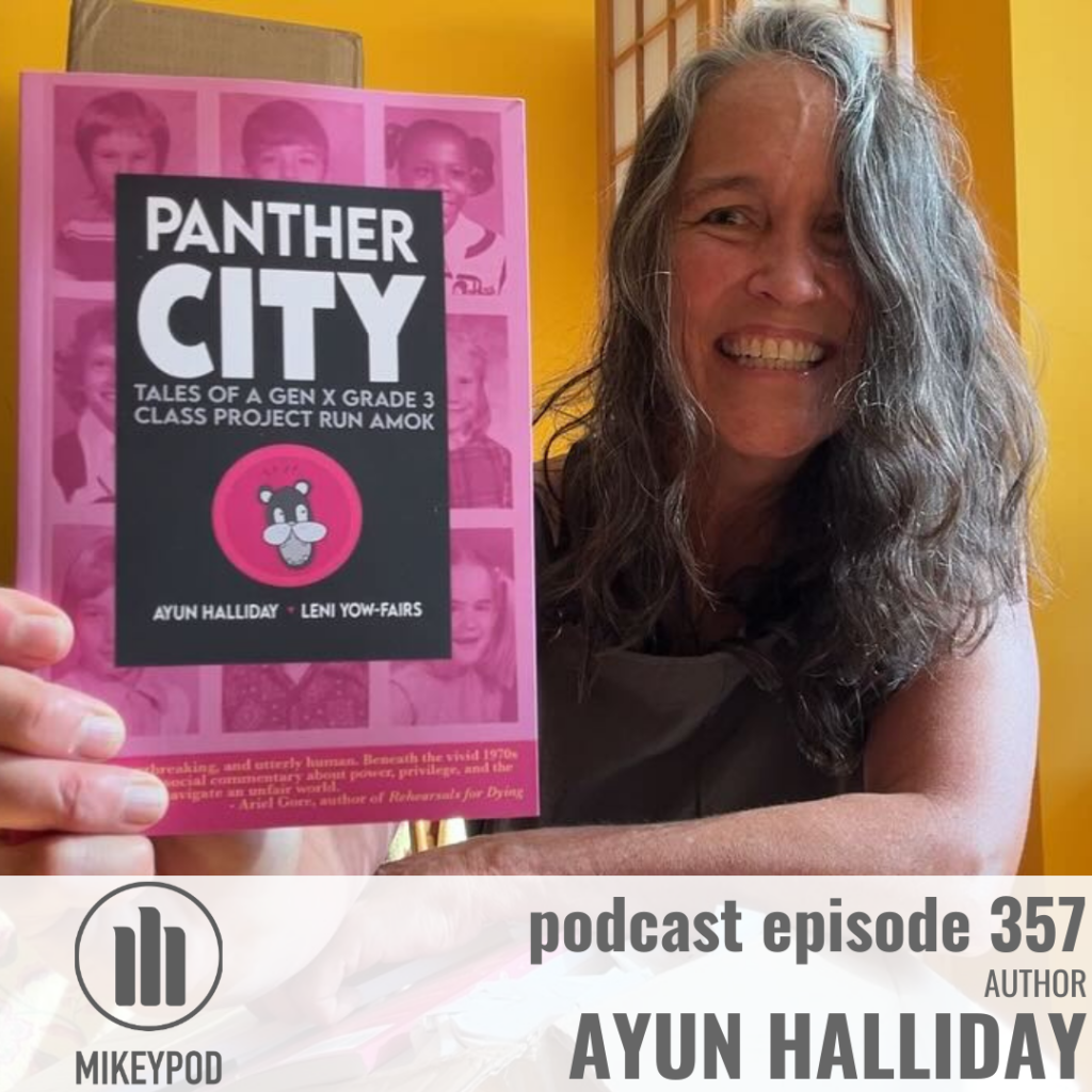 A woman with long dark brown and gray hair smiles while holding up a pink book titled “Panther City: Tales of a Gen X Grade 3 Class Project Run Amok.” The background shows a warmly lit yellow room with a wooden screen. Text and the MikeyPod logo are overlaid along the bottom to promote a podcast episode.