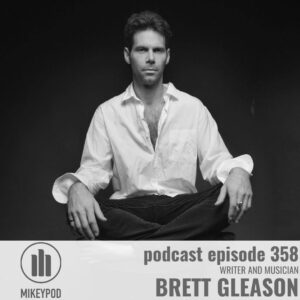 Black and white portrait photograph of Brett Gleason, a man in a white button-up shirt with rolled sleeves and dark pants, seated against a dark background. The MikeyPod logo appears in the lower left, with text reading "podcast episode 358, Writer and Musician, Brett Gleason" on the right side