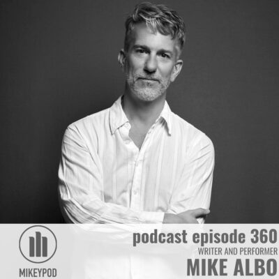 Black and white portrait of Mike Albo, a man with short salt-and-pepper hair and beard wearing a light cream button-up shirt, arms crossed, against a dark gray background. The MikeyPod logo appears in the lower left, with text reading "podcast episode 360, Writer and Performer, Mike Albo" on the right.